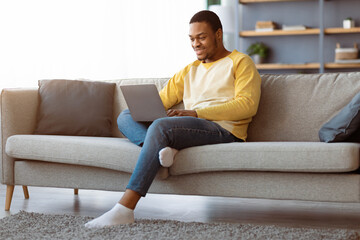 Black guy spending time at home, using modern laptop, sitting on couch, surfing on Internet, copy...