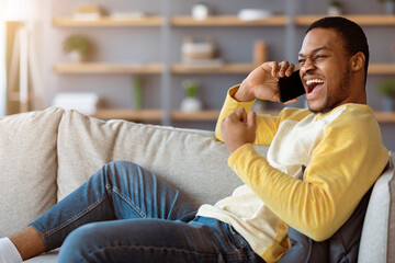 Fototapeta premium Communication concept. Emotional african american man sitting on sofa in living room, having conversation on mobile phone, got good news, screaming, gesturing and looking at copy space, home interior