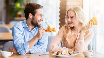 Naklejka premium Positive young couple eating healthy breakfast together at coffee shop. Handsome millennial man having dinner with his girlfriend at cafe, enjoying yummy meal, spending happy time with beloved woman