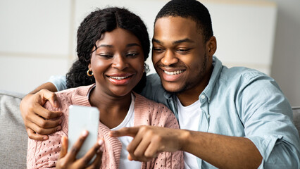 Happy black wife showing her husband photos on smartphone while sitting together on couch in living room, closeup. African american smiling couple using mobile phone at home, playing game, copy space