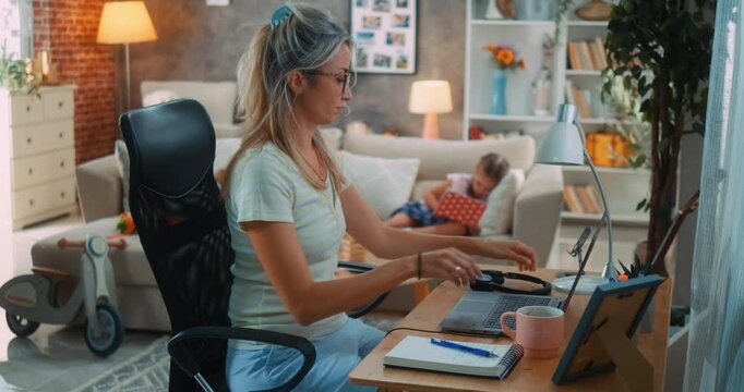 Busy mother prepares for remote work video call with headphones, unaware of her sad daughter playing alone and seeking attention in the background at home in living room.