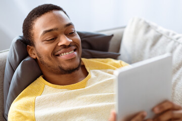 Closeup of smiling black guy using digital tablet while laying on couch at home, copy space....