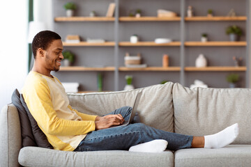 Side view of happy african american guy enjoying weekend at home, sitting on couch in cozy living...