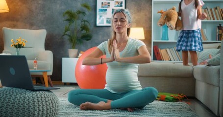 Pregnant mother trying to meditate online with laptop at home, while her playful daughter jumps on the bed and throws a teddy bear, interrupting peaceful moment of relaxation. - Powered by Adobe