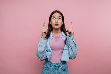 Young woman in casual outfit pointing upwards against pink background.
