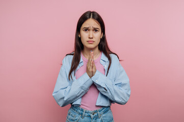 Fototapeta premium Young woman with long hair in casual clothes, hands in prayer position, looking worried, standing against a pink background.