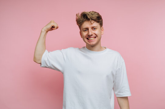 Smiling young man in white t-shirt flexing arm on pink background.