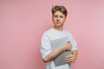 Young man in white t-shirt holding a laptop against a pink background.