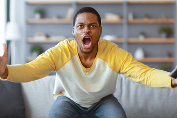 Furious black guy watching football game on TV, yelling and gesturing with remote in his hand,...
