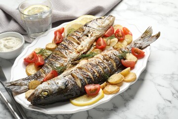 Tasty roasted seabass fish served with potatoes, tomatoes, sauce and lemon on white marble table, closeup. Homemade seafood dish
