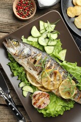 Tasty roasted seabass fish served with vegetables and lemon on wooden table, flat lay. Homemade seafood dish