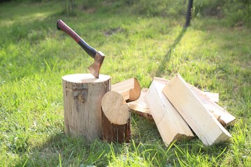 Metal axe, chopped firewood and wooden log on green grass outdoors