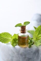 Bottle of aromatic essential oil and herbs in marble mortar against white background, closeup