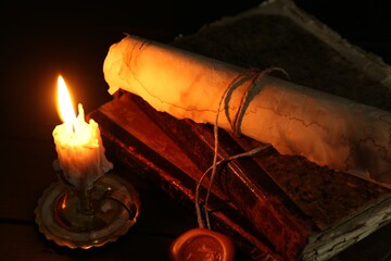 Old rolled scroll, burning candle and books on black wooden table, closeup