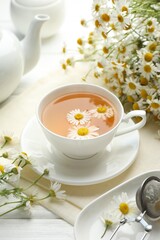 Tasty chamomile tea, infuser and flowers on white wooden table, closeup