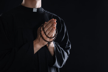 Priest with rosary praying on black background, closeup. Space for text