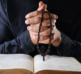 Priest with rosary and Holy Bible praying on black background, closeup