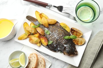 Tasty roasted flounder fish served with potatoes, basil and lemon slices on white marble table, flat lay. Homemade seafood dish