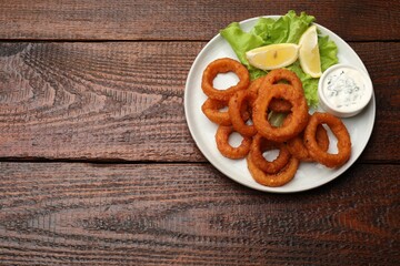 Fried squid rings, lemon, lettuce and sauce on wooden table, top view. Space for text