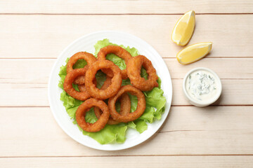 Fried squid rings, lemon, lettuce and sauce on light wooden table, flat lay