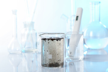 Glass beaker with dirty water, other glassware and digital pH meter on mirror surface, closeup