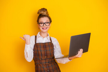 Young female student with a laptop on a yellow background showing a pointing gesture wearing...