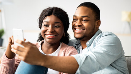 Cheerful loving african american young couple taking selfie while spending time together at home, using smartphone, closeup. Happy black man and woman using mobile phone, choosing something