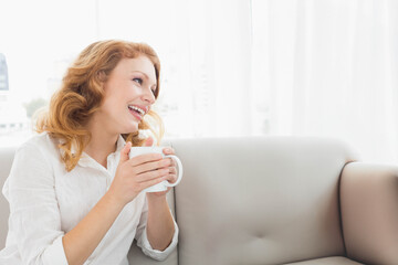 Woman holding white ceramic mug and smiling while sitting on beige sofa in bright living room
