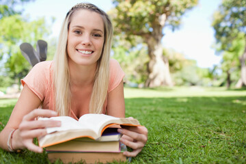 Fototapeta premium Female student lying on park grass reading open book on stacked books under bright sunlight