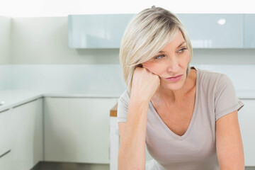 Mature woman sitting at glossy countertop leaning chin on hand gazing left in kitchen, copy space
