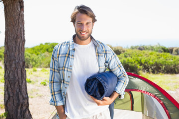 Man holding rolled-up sleeping bag while standing in woodland campsite near dome tent