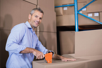 Middle-aged man using orange tape dispenser sealing cardboard boxes at warehouse with blue shelving