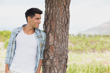 Man leaning against tree trunk in field amid low shrubs overlooking mountain ridge