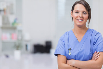 Female nurse in blue scrubs smiling while standing wearing ID badge in examination room, copy space