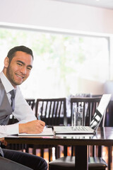 Asian man writing notes in notebook at cafe table with open laptop, water glass, copy space