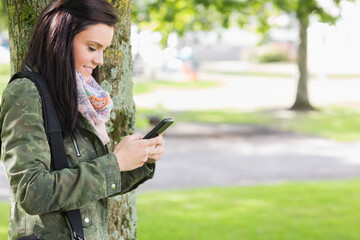 Female leaning against tree trunk in park, checking smartphone and carrying black bag, copy space