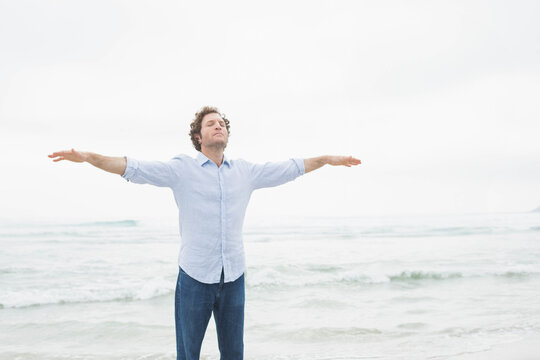 Middle-aged man extending arms while standing waist-deep at beach shoreline under overcast sky