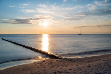 Ein Segelboot am Horizont der Ostsee während eines Sonnenuntergangs und im Vordergrund eine Buhne