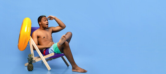 Millennial black guy in swimwear sitting in lounge chair, holding hand above his eyes, looking into...