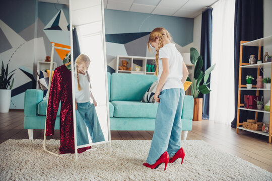 Young girl trying on oversized red high heels while standing in front of a mirror in a cozy modern living room
