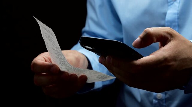 Businessman checking a receipt for purchases on a black background.
Paper receipt for non-payment in the hands of a businessman.
Man scanning information on a receipt