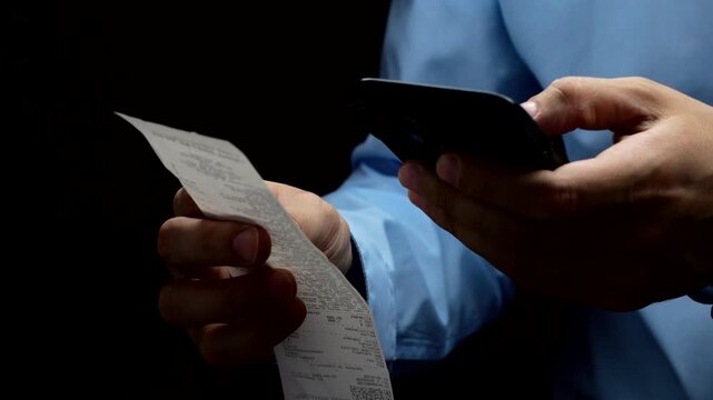 Businessman checking a receipt for purchases on a black background.
Paper receipt for non-payment in the hands of a businessman.
Man scanning information on a receipt