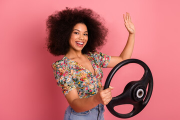 Cheerful young woman posing with a steering wheel on a vibrant pink background, showcasing joy,...
