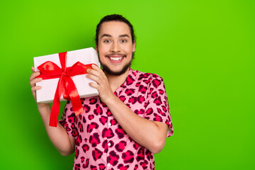 Cheerful young man holding a wrapped gift box with a red ribbon, wearing a pink leopard-print...