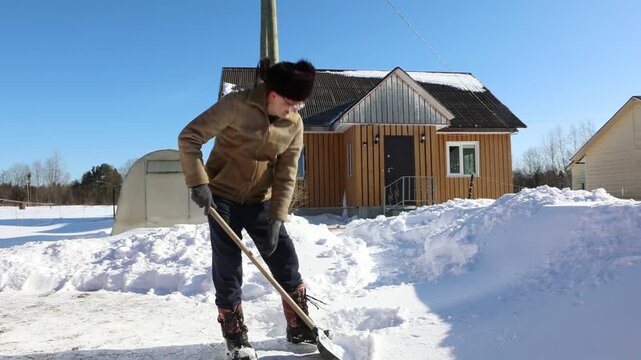 Lifting snow using snow shovel by individual in insulated coat and fur hat near wooden farm house with snow piles around.