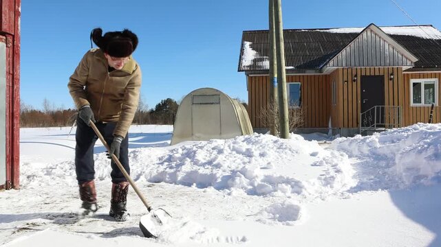 Shoveling snow with plastic shovel by man in fur hat and warm coat in front of red wooden house under bright winter sun.