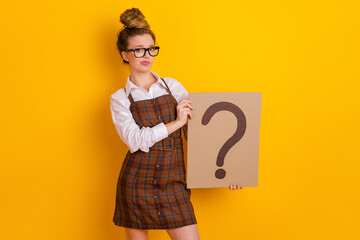 Adorable woman in plaid dress holding a question mark sign on a yellow background, expressing...