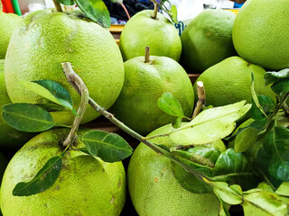 A close-up of a pile of fresh, green pomelo fruit with stems and leaves, ready for sale at a market.