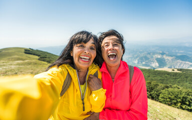 Two senior women taking selfie on top of the mountain - Happy aged female friends having fun on trekking excursion - Sport and tecnology life style concept