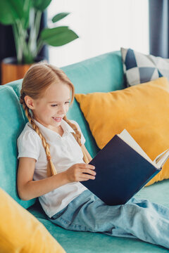 Young girl relaxing at home while reading a book on a cozy teal couch with bright yellow pillows